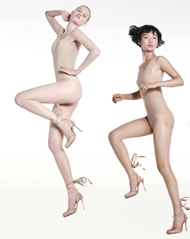Two fashion models pose dynamically in nude bodysuits and matching heels against a clean white backdrop, highlighting form and movement.