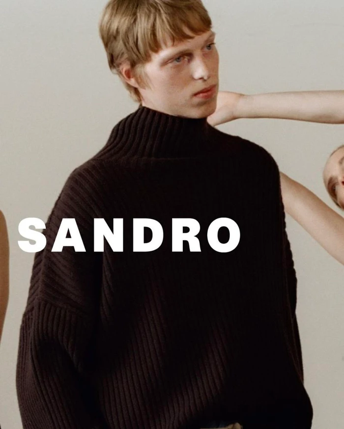 Male model in a dark ribbed knit turtleneck sweater posing against a neutral backdrop with hands reaching toward him.