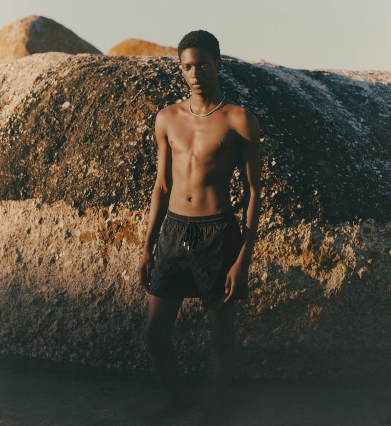 Young adult male standing in water on a beach, wearing shorts, posing against a large rock formation on a sunny day.