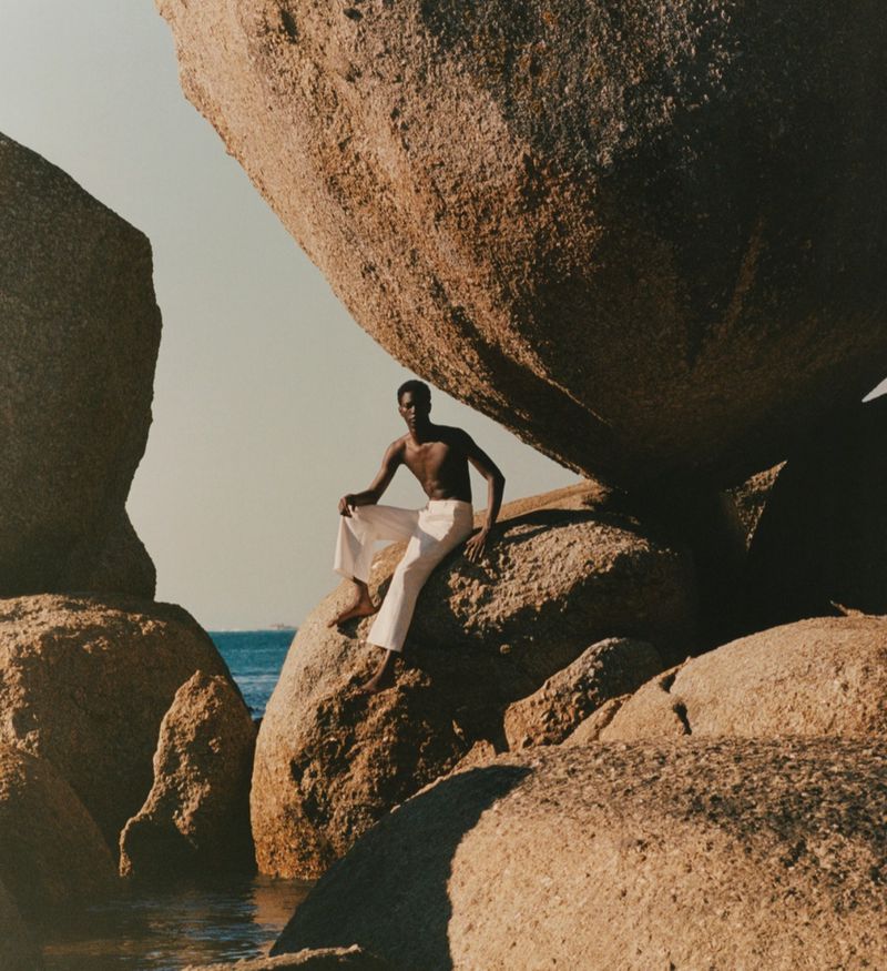 An adult male sits on a rock formation at the beach wearing white pants in a sunny outdoor setting with blue water visible.