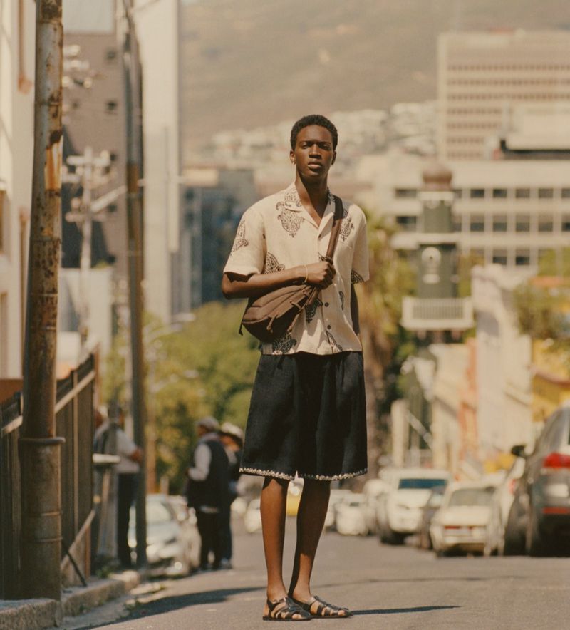 A young adult man stands on a city street wearing a patterned button-down shirt, dark shorts, sandals, and a cross-body bag in daytime.