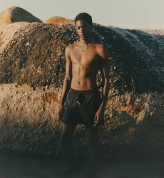 Shirtless black male model wearing dark swim trunks standing against large rocky boulders outdoors