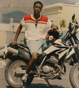 Model Limamu Mbaye seated on a motorcycle holding a helmet for the Mr Porter x Loewe High Summer Campaign, photographed by Quentin De Briey