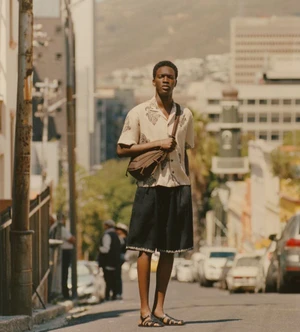 Black man standing on a city street wearing patterned short-sleeve shirt, dark shorts, and carrying a brown crossbody bag.
