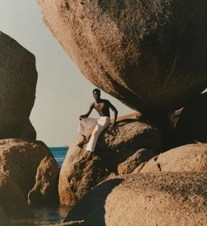 Shirtless man wearing white trousers seated on large rock formation near ocean water by Quentin De Briey