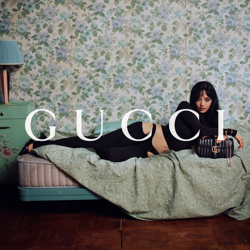 Ningning reclines on a bed in a black cut-out outfit next to a black quilted handbag against floral wallpaper.