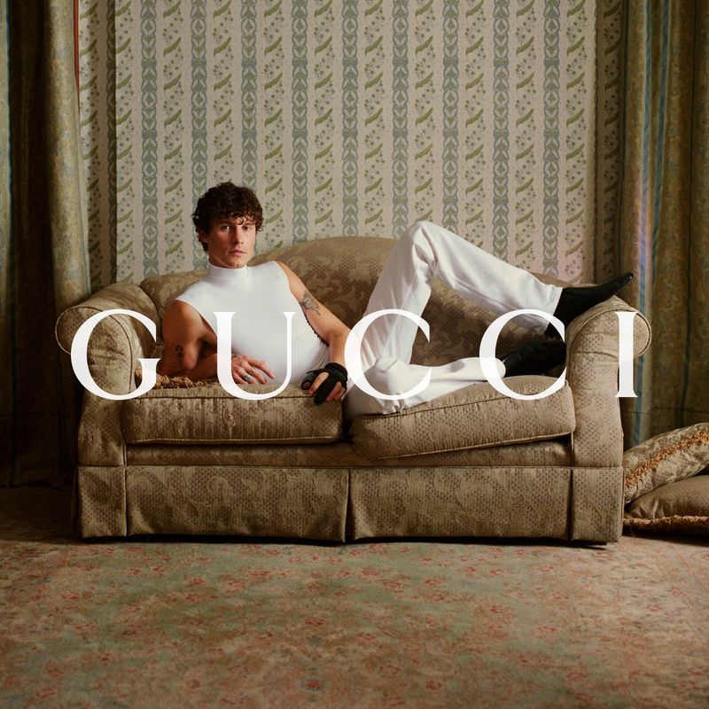 Shawn Mendes reclining on a beige patterned vintage sofa in a sleeveless white top and white trousers for Gucci.