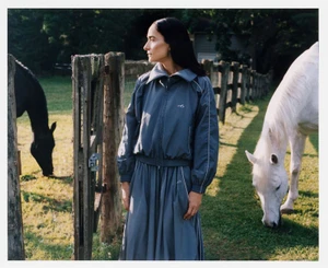 A woman in a slate blue track jacket and skirt stands outdoors by a rustic wooden fence with a black horse and a white horse.
