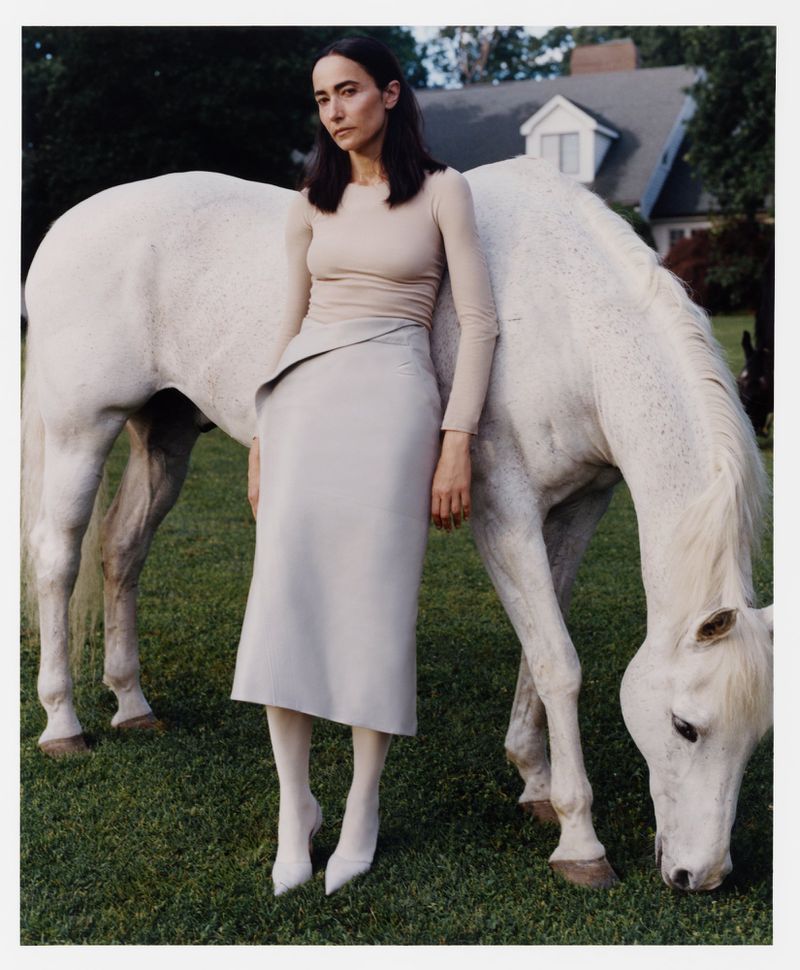 Model in a beige top and pale grey midi skirt standing beside a grazing white horse on a lawn with a house in the background.