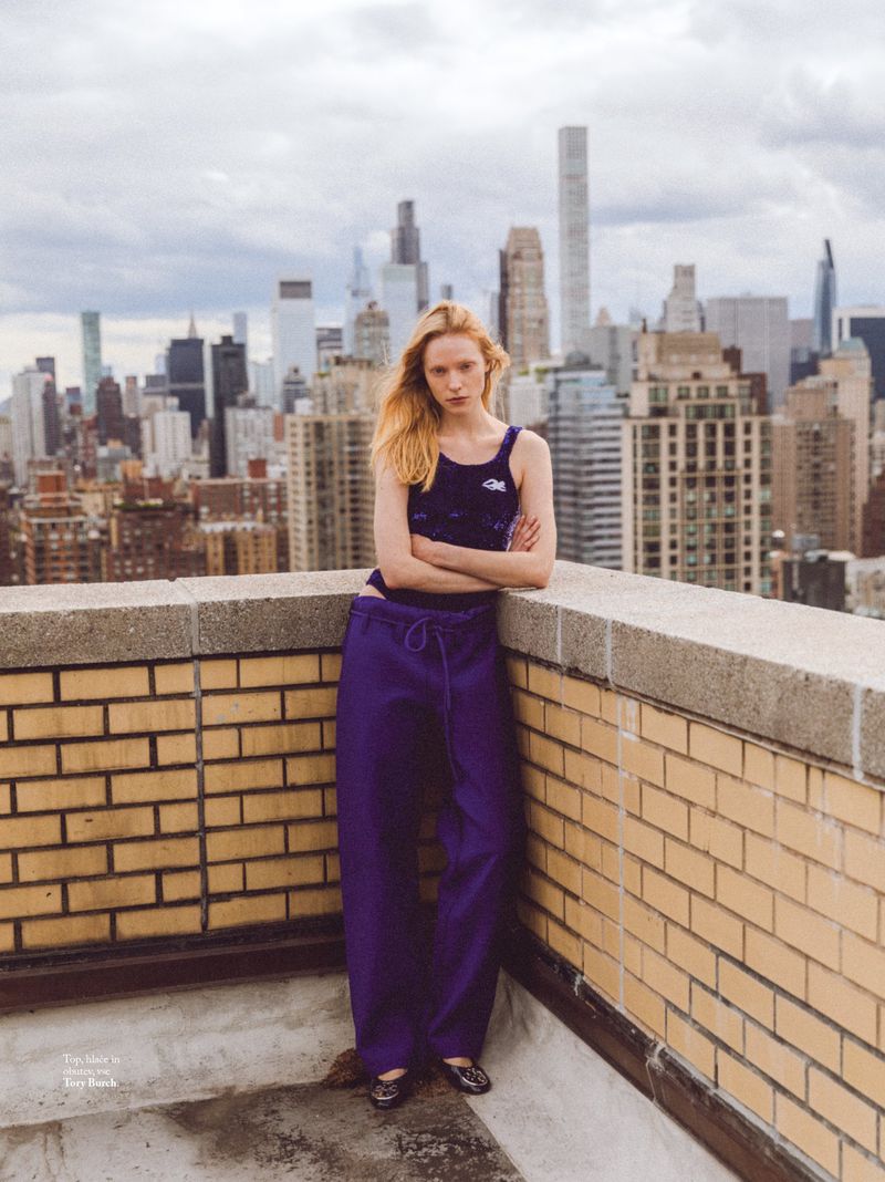 Young woman in a purple top and loose-fitting pants poses on a rooftop overlooking a cityscape on an overcast day.