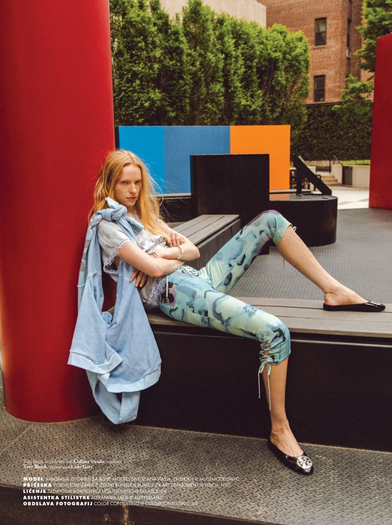 Fashion editorial shot of a blonde model reclining on a bench outdoors, wearing a patterned top and leggings, and a light denim jacket.