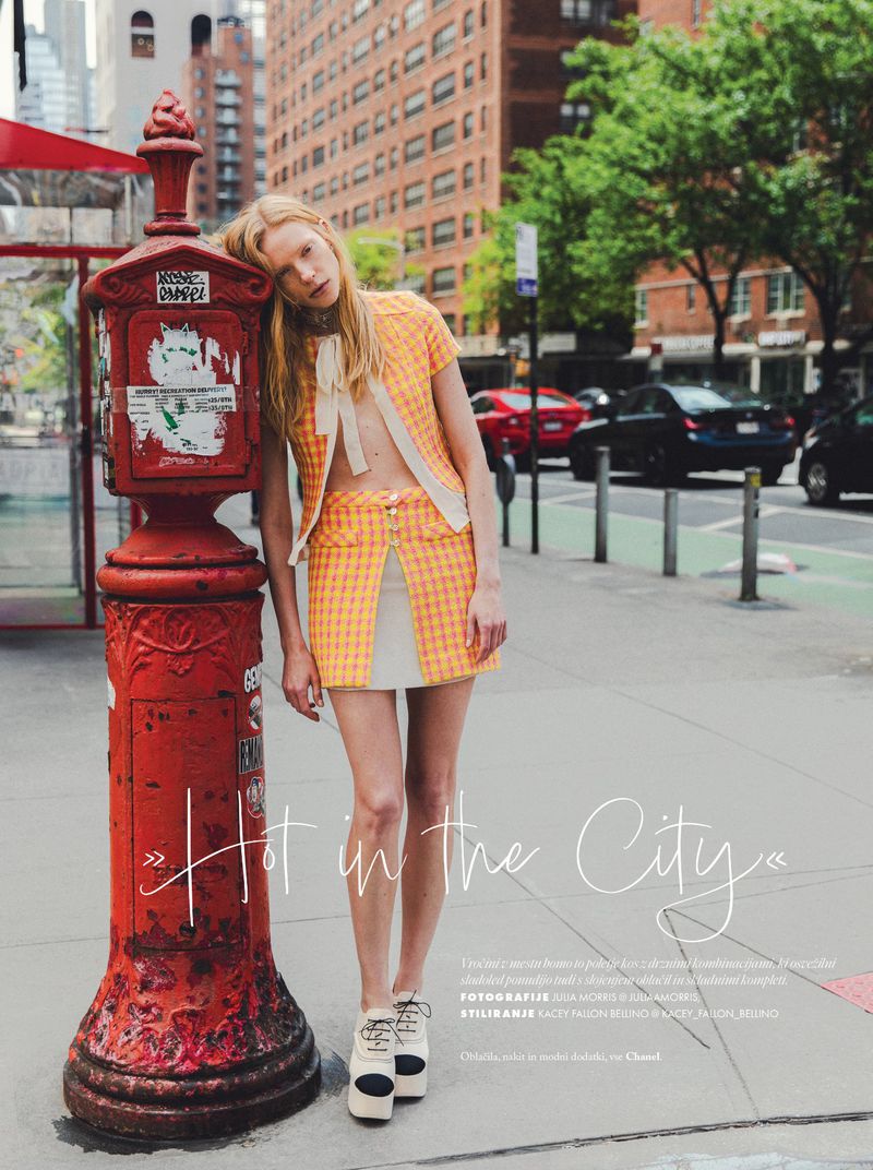 Fashion editorial shot of a blonde woman posing in a yellow and white skirt and jacket set, leaning against a red vintage post box on a city street.