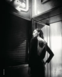 Model Noa Kaiser viewed from behind in a backless black long sleeve dress with a floral corsage ornament under glowing neon lighting.