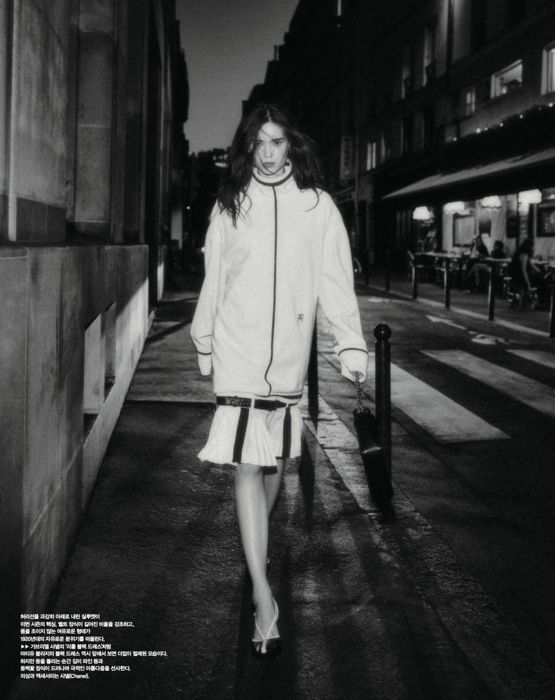 Model Noa Kaiser walking along a city street at night in a white drop-waist dress with black trim and carrying a handbag.