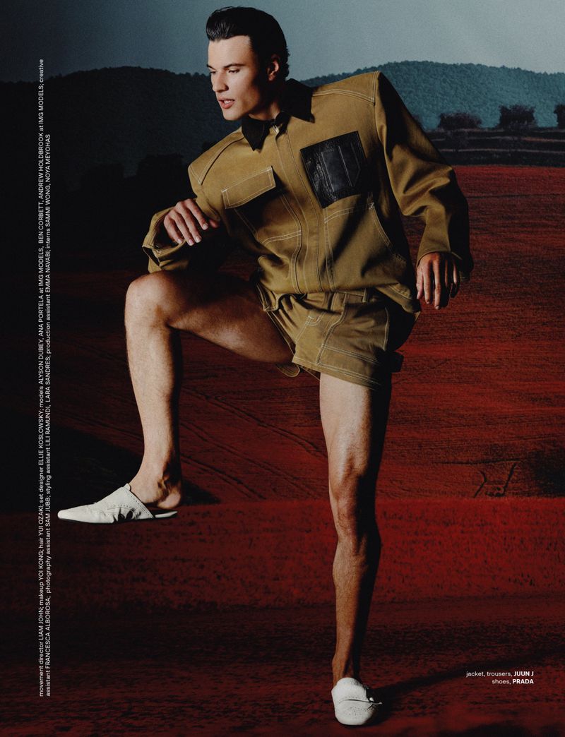 A stylish adult male poses in a tan shirt and shorts with white shoes, leg raised, against a background of reddish-brown and distant mountains.