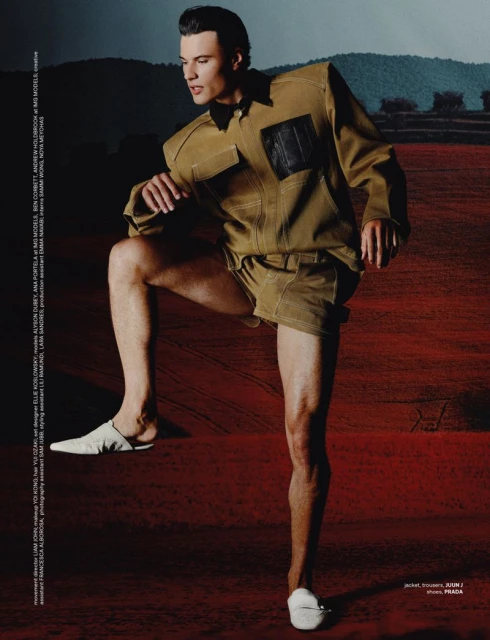 A stylish adult male poses in a tan shirt and shorts with white shoes, leg raised, against a background of reddish-brown and distant mountains.