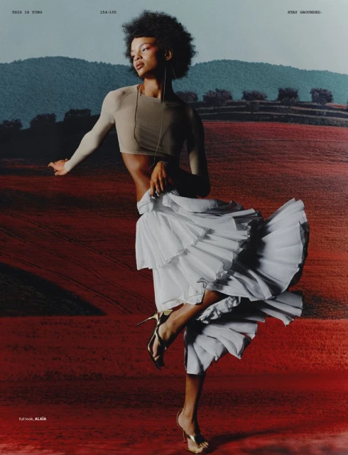 Fashion editorial shot of a Black female model posing in a flowing white skirt and gold heels, set against a landscape backdrop.