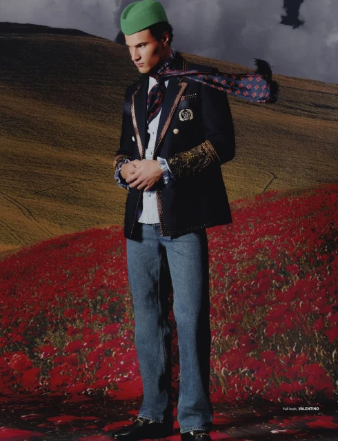 A man stands in a poppy field wearing a green hat, a dark jacket with gold trim, a patterned scarf, and jeans in a landscape setting.