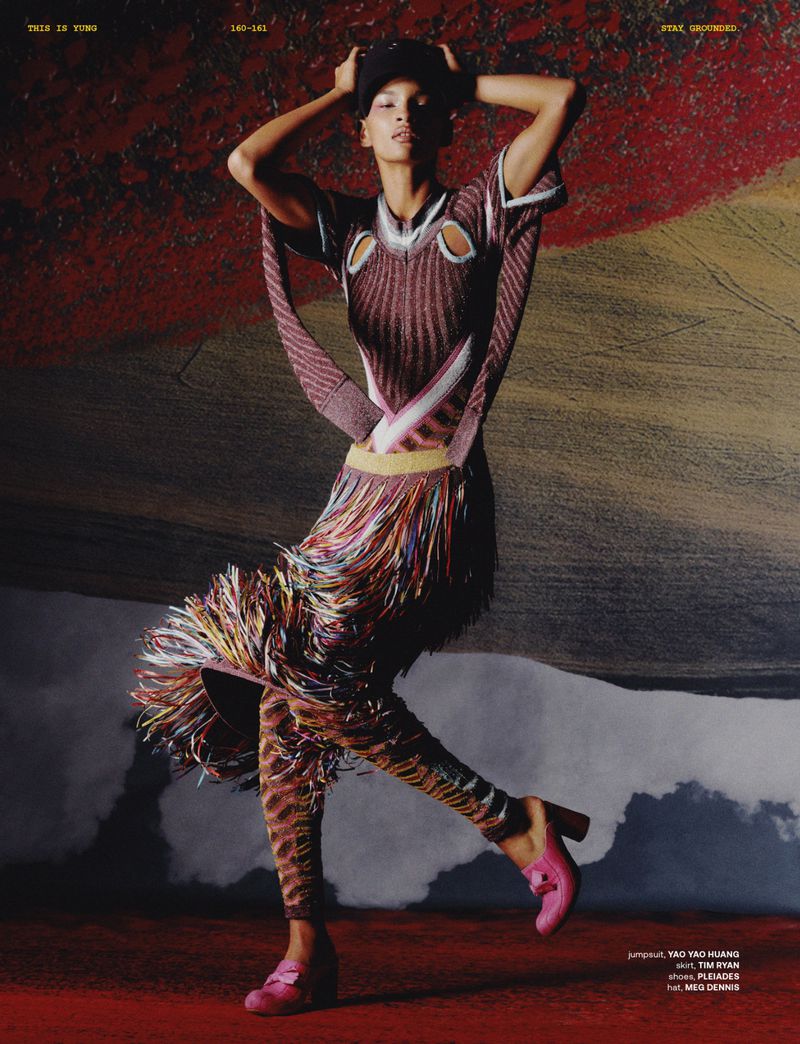 Editorial fashion shot of a slender female model in a patterned tunic and leggings, posing against a textured backdrop with pink heels and cap.