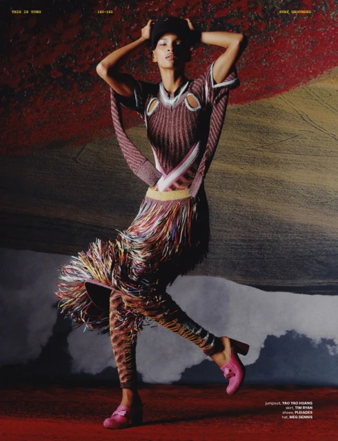 Editorial fashion shot of a slender female model in a patterned tunic and leggings, posing against a textured backdrop with pink heels and cap.