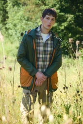 Model Ryder Forsyth wearing a green and orange utility jacket and plaid shirt standing in a tall grass field.