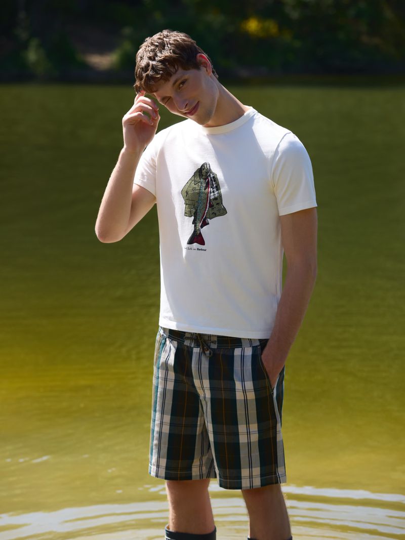 Model Ryder Forsyth standing in lake water wearing a white graphic t-shirt and plaid shorts, touching his hair.