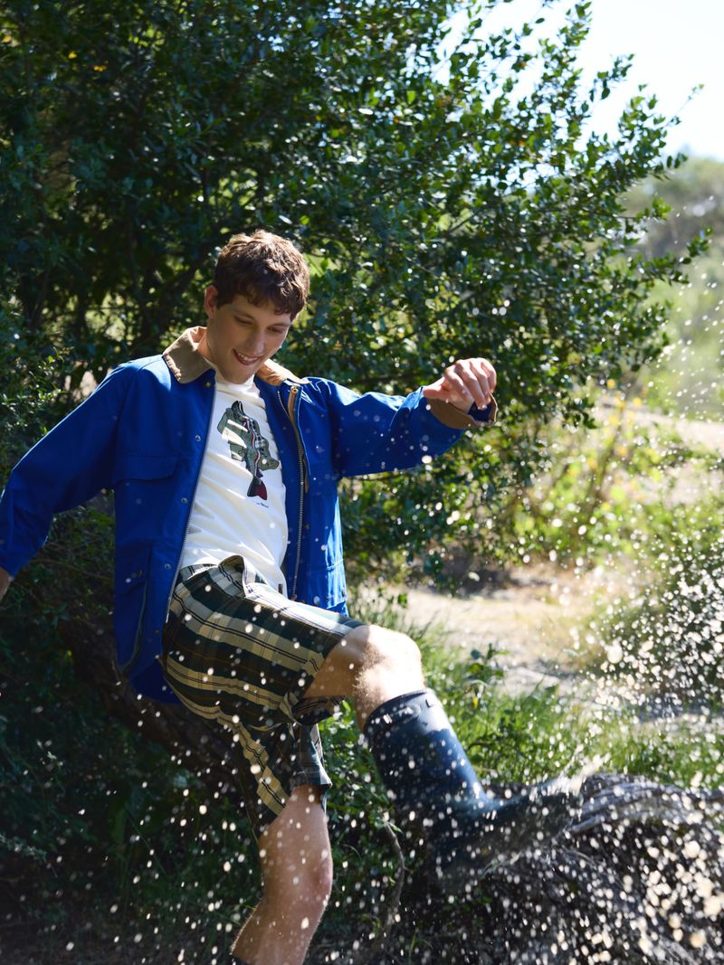 Man in a bright blue jacket and plaid shorts kicks water outdoors, sending water droplets flying against green foliage.