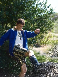 Man in a bright blue jacket and plaid shorts kicks water outdoors, sending water droplets flying against green foliage.