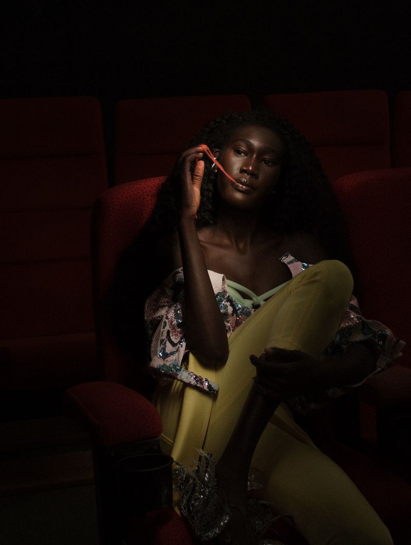 Black woman seated in a red velvet cinema chair stretching pink bubblegum from her mouth under dramatic low lighting.