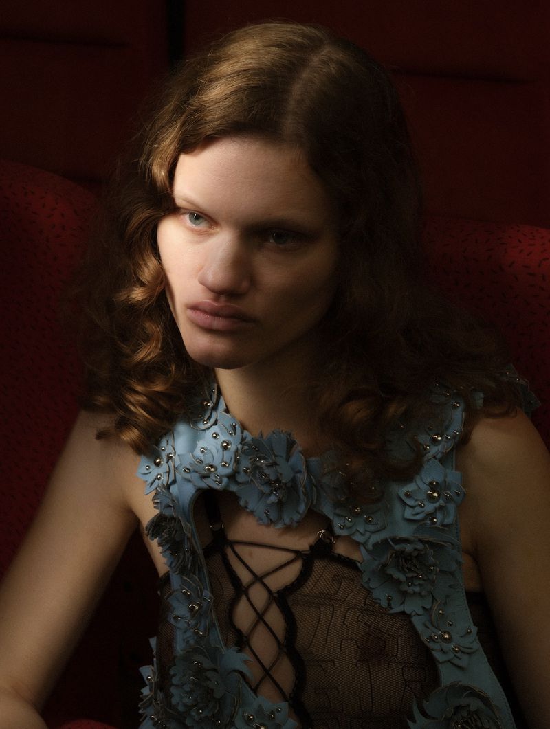 Model with wavy brown hair wearing a light blue floral cutout top over black sheer lace in red velvet seating.