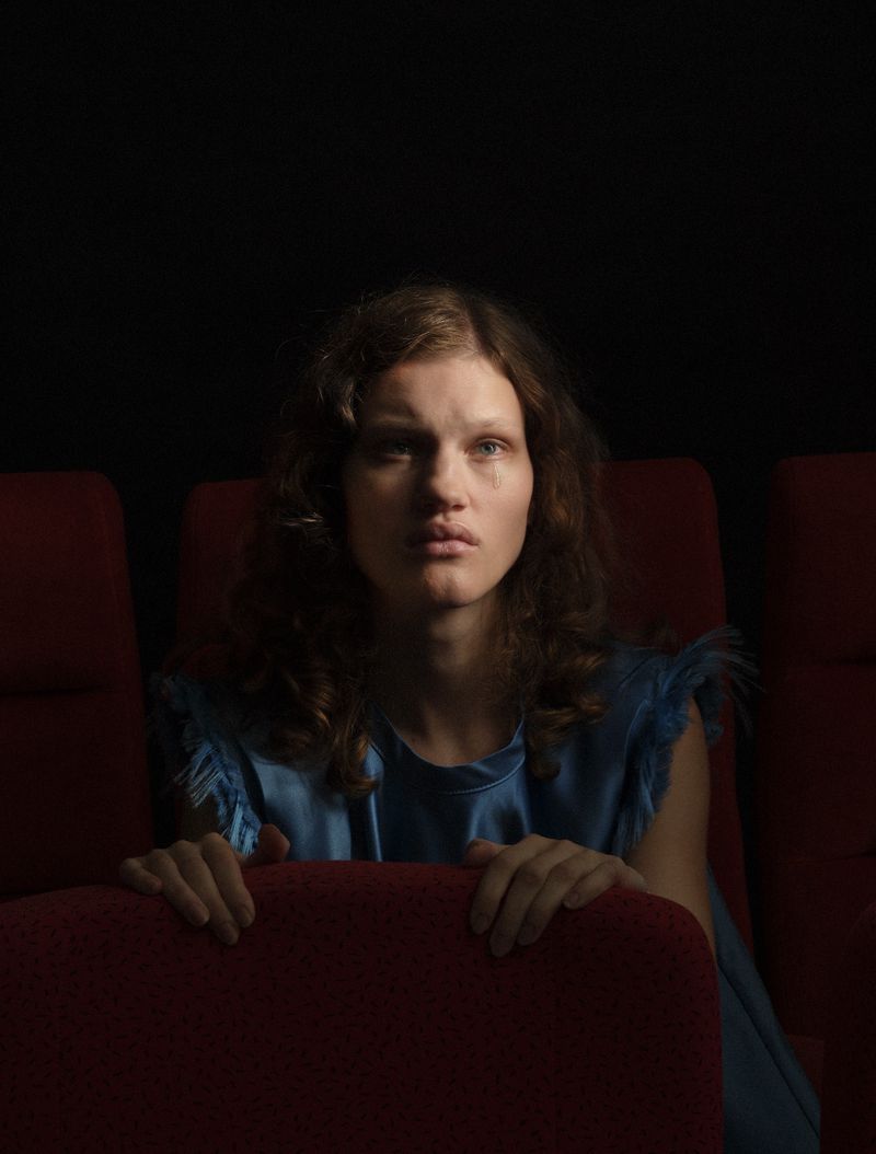Model with wavy brown hair wearing a blue ruffled satin top seated behind dark red theatre chairs.