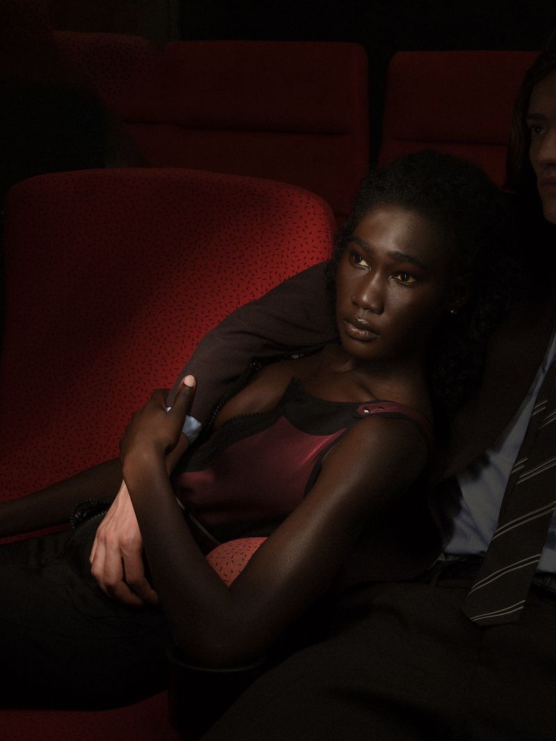A Black woman wearing a dark sleeveless top sits in dim red theater seats with an arm resting over her shoulder.