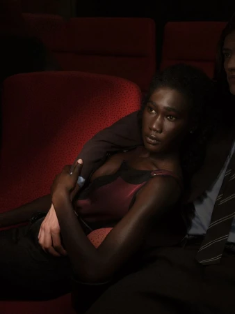 A Black woman wearing a dark sleeveless top sits in dim red theater seats with an arm resting over her shoulder.