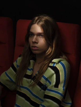Model with long hair wearing a green striped collared knit shirt seated in a red theatre chair in low lighting.