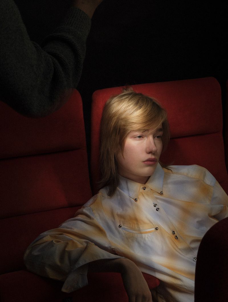 A model with blonde hair wearing a patterned shirt reclines in red velvet cinema seats in dramatic spotlight.