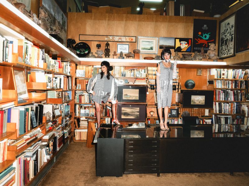 Two models in patterned grey dresses pose inside a room filled with bookshelves, with one standing atop a black executive desk.