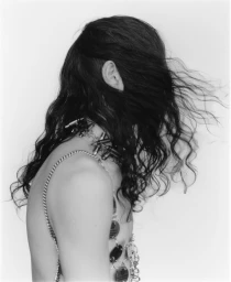 Side profile of a model with long dark wavy hair obscuring the face, wearing metal chain accessories against a white backdrop.