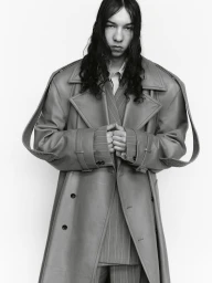 Model with long dark wavy hair wearing an oversized trench coat and pinstripe suit against a white studio background.