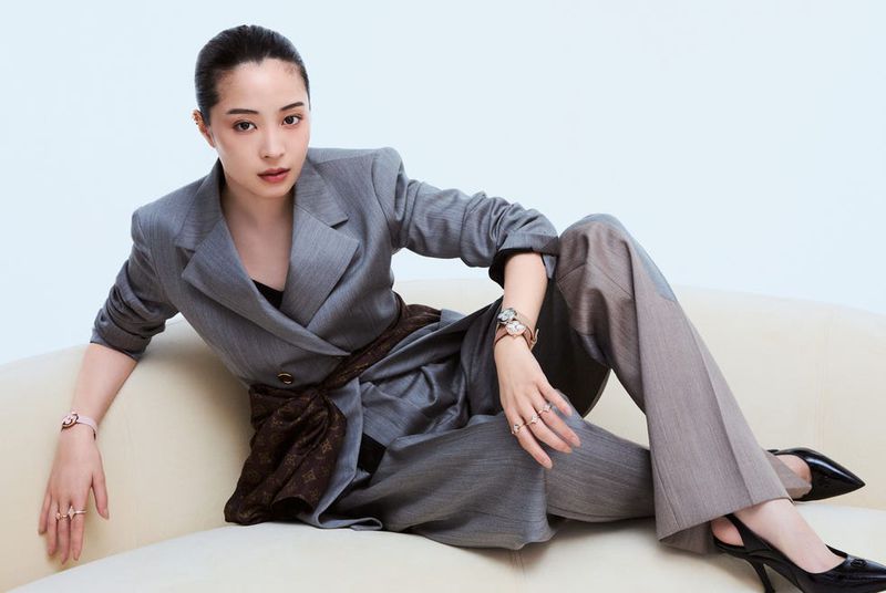 Suzu Hirose reclining on a cream curved couch in a grey suit with a monogram sash and black heels in a studio setting.