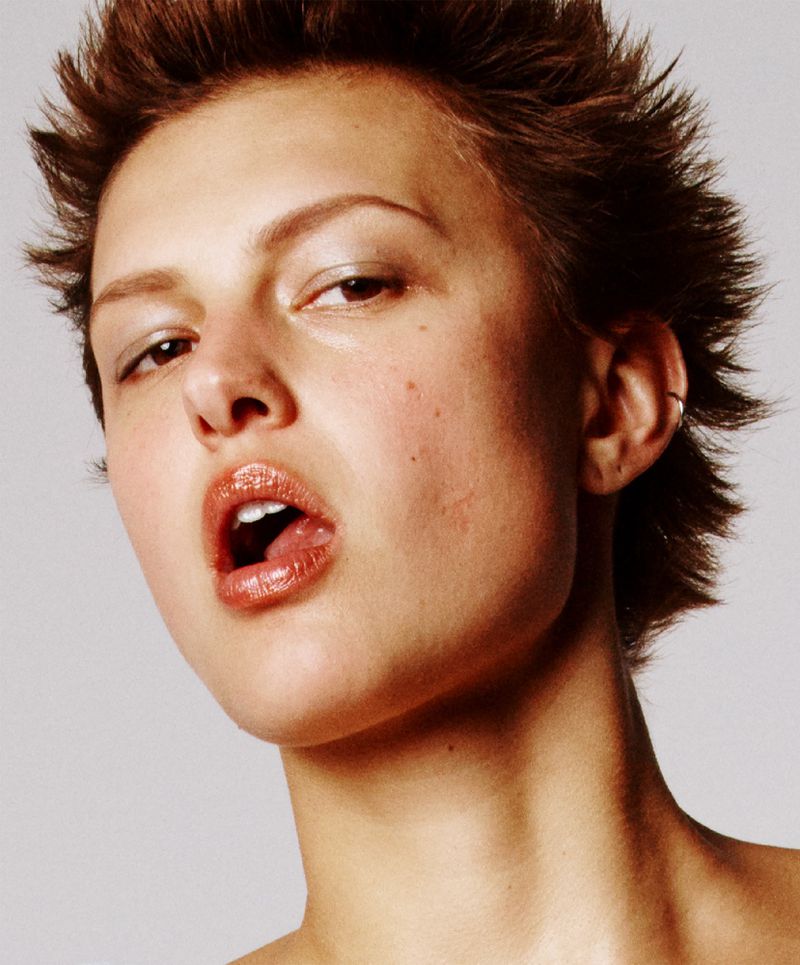 Close-up portrait of a young woman with short brown hair, glossy lips, and fair skin, looking up with her mouth slightly open.