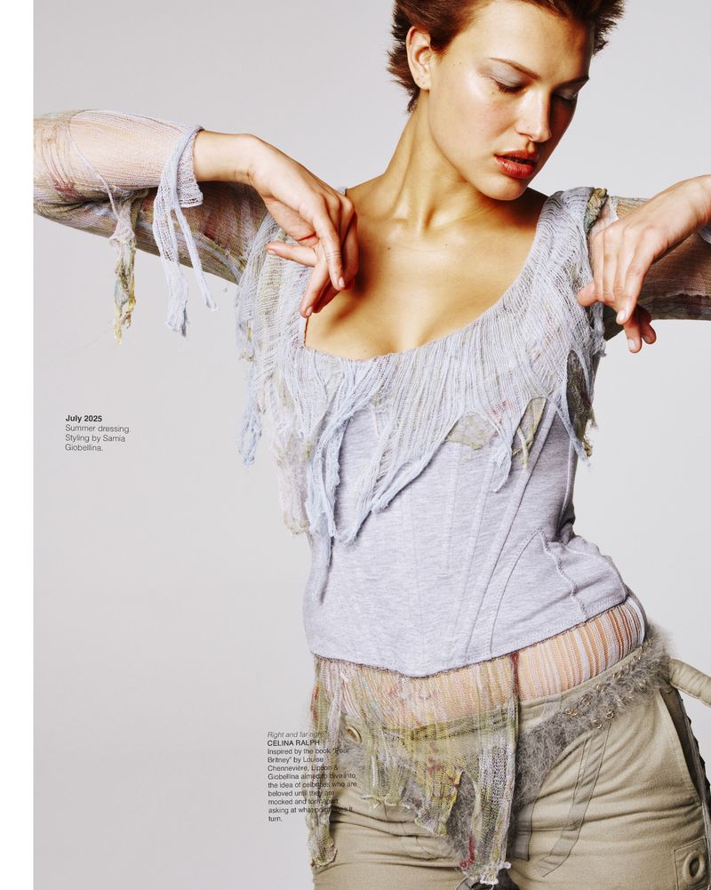 Woman poses in fashion shot wearing gray top, tan pants with distressed and draped fabrics, set against a plain background.