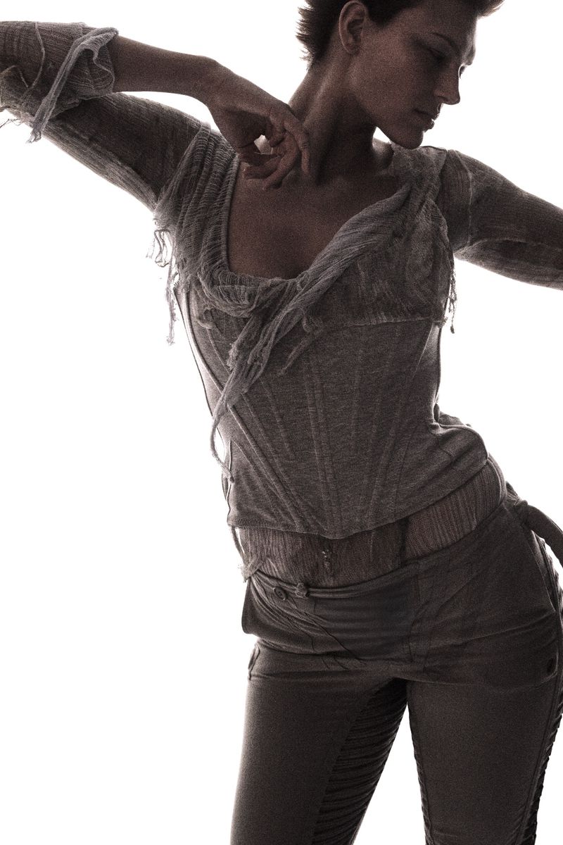 Fashion shot of a slender adult woman in a light-colored, distressed top and dark pants, striking a dynamic pose against a white backdrop.