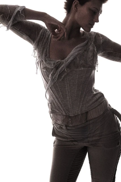 Fashion shot of a slender adult woman in a light-colored, distressed top and dark pants, striking a dynamic pose against a white backdrop.