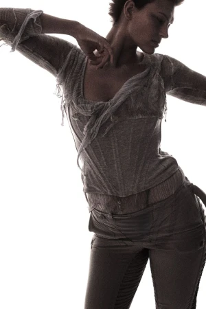 Fashion shot of a slender adult woman in a light-colored, distressed top and dark pants, striking a dynamic pose against a white backdrop.