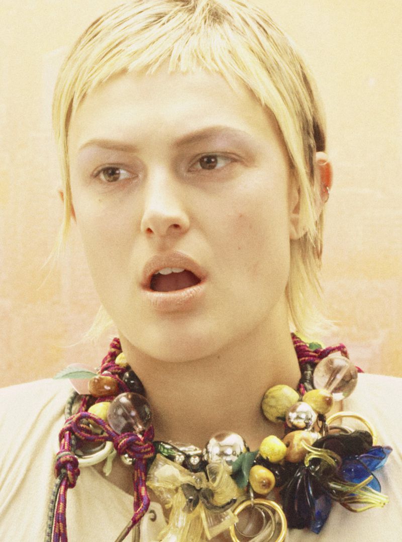 Close-up shot of a young adult woman with light blonde hair, wearing a colorful statement necklace with various beads and charms.