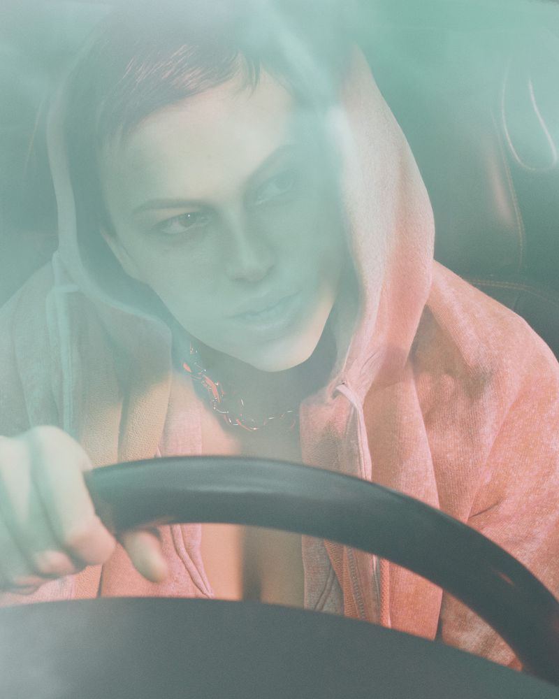 An androgynous person in a hooded sweatshirt driving a car, with a view of the steering wheel and dashboard from the back seat.