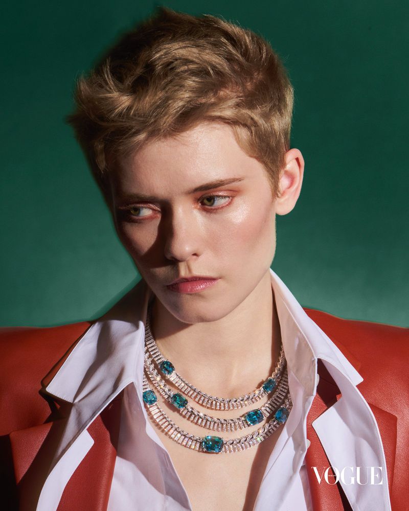 Model Maike Inga with short blonde hair wearing a layered diamond and blue gemstone necklace and rust blazer on a green background.