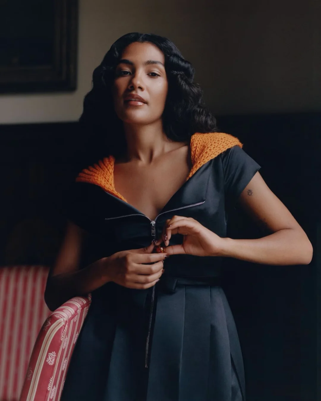 Olivia Dean wearing a dark zip-front dress with an orange knit collar, posing beside a striped armchair in soft lighting.