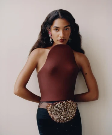 Olivia Dean wearing a sculptural burgundy molded top and black skirt with hands behind her back against an off-white backdrop.