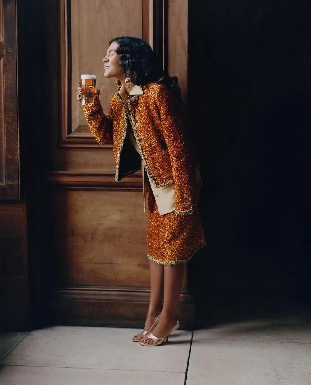 Olivia Dean posing in a shimmering copper tweed skirt suit while holding up a glass of beer against wooden wall panelling.